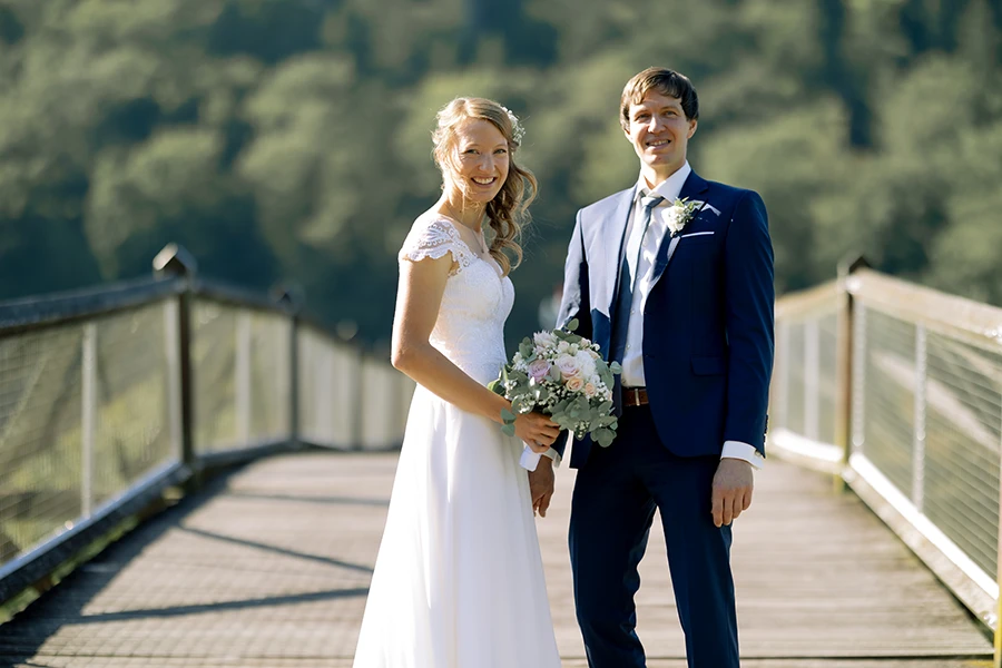 Das Brautpaar steht auf einer Brücke und lächelt in die Kamera.